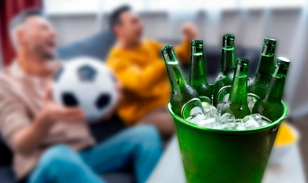 Balde com garrafas de cerveja verdes cheias de gelo em primeiro plano, enquanto duas pessoas assistem a um jogo de futebol no sofá ao fundo, representando bebidas geladas no verão.