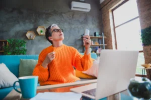 Pessoa trabalhando em home office em um ambiente residencial, sentada no sofá com notebook aberto, usando controle remoto para ajustar o ar-condicionado na parede, em um espaço bem iluminado e organizado.