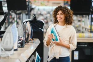 Mulher sorridente escolhendo um novo ferro a vapor em uma loja de eletrodomésticos, comparando diferentes modelos em exibição