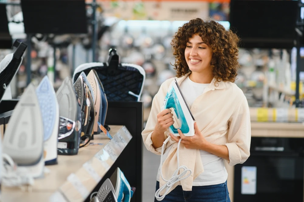 Mulher sorridente escolhendo um novo ferro a vapor em uma loja de eletrodomésticos, comparando diferentes modelos em exibição