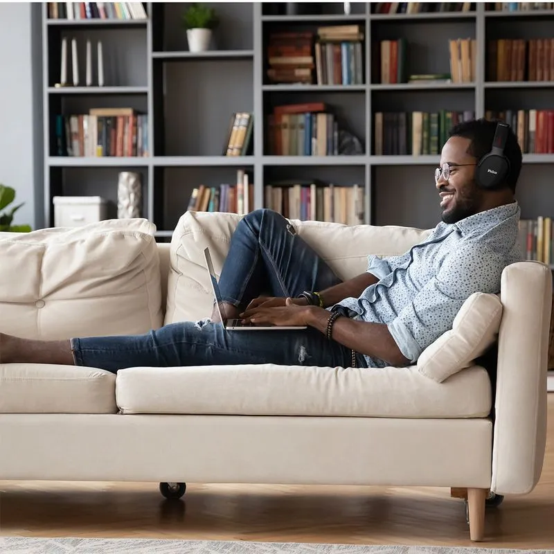 Homem sorridente sentado de forma relaxada num sofá bege, utilizando um portátil e fones de ouvido headphone Philco pretos, ilustrando o uso de tecnologia para lazer ou home office em ambiente doméstico.